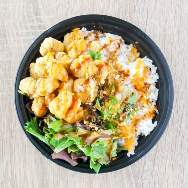 Top-down view of a black bowl filled with popcorn chicken, white rice, green salad, and sauces.