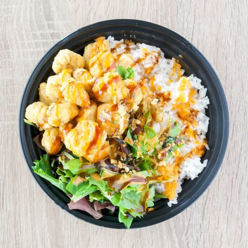 Top-down view of a black bowl filled with popcorn chicken, white rice, green salad, and sauces.