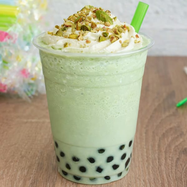Green bubble tea with whipped cream, crushed pistachios, and a green straw on a wooden table.